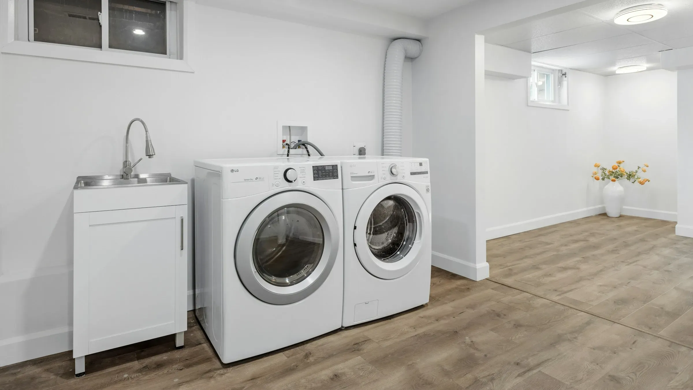 A washer and dryer side by side in a plain utility room, with a sink beside them and hose connections on the wall behind.